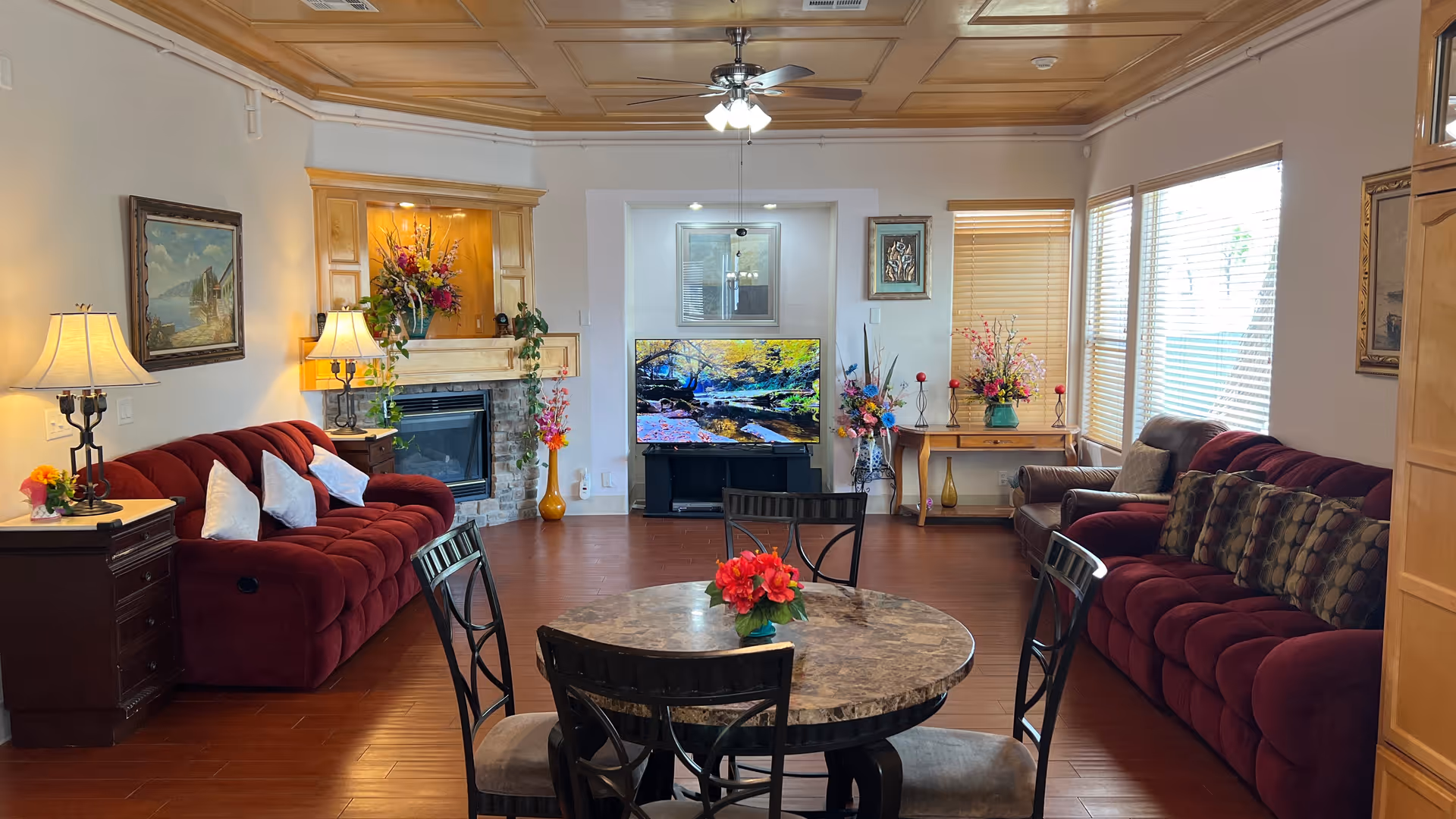 A cozy senior living room with two red sofas, a round marble-top table with four chairs, a fireplace with a floral arrangement above it, a flat-screen TV displaying a nature scene, and large windows with blinds letting in natural light. The room is decorated with paintings, lamps, and various flower vases.