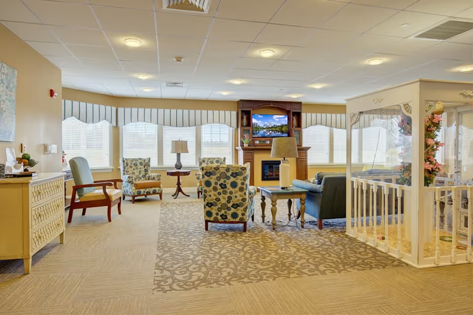 Bright senior living common room with upholstered chairs and a sofa arranged around a fireplace and TV, large windows along the back wall, lamps, and a small decorative enclosed seating area.