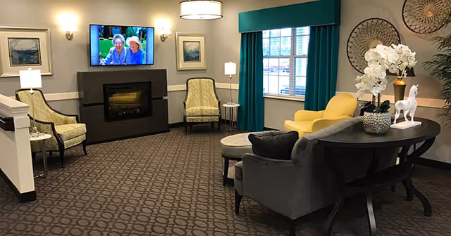 A cozy living room area with patterned carpet, two green armchairs, a yellow armchair, a gray sofa, and a black console table with white orchids and a decorative horse figurine. A flat-screen TV is mounted above a fireplace, flanked by two wall lamps and framed artwork. A window with teal curtains lets in natural light.