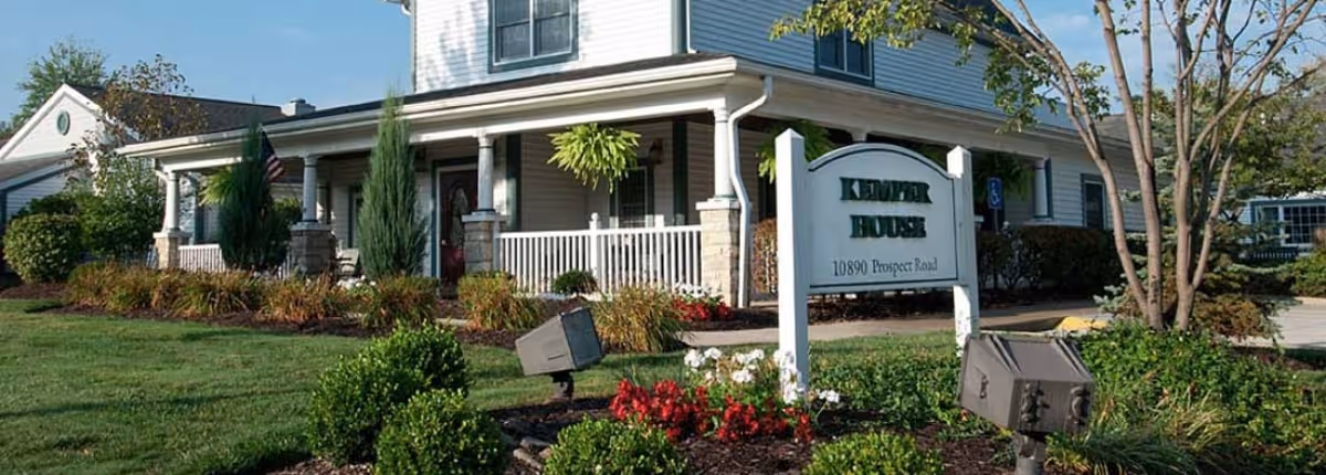 Front exterior of the Kemper House building with a porch, white sign, and landscaped lawn and flowerbeds.