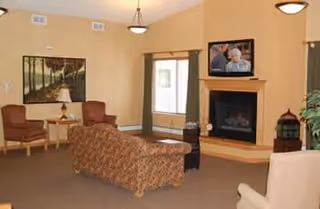 A cozy senior living community lounge with a patterned sofa, two brown armchairs, a beige armchair, a fireplace with a TV mounted above it, a large window with curtains, a framed landscape painting on the wall, and soft overhead lighting.