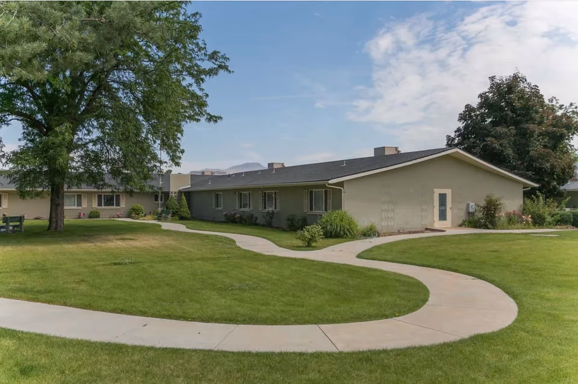 A single-story building with beige walls and a dark roof surrounded by a well-maintained green lawn and a curved concrete walkway. There are several windows along the building and a large tree on the left side. The sky is partly cloudy with patches of blue.