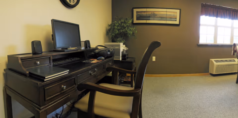 A dark wood desk with a computer and chair in a carpeted room with a window, wall art, and an air conditioner unit.