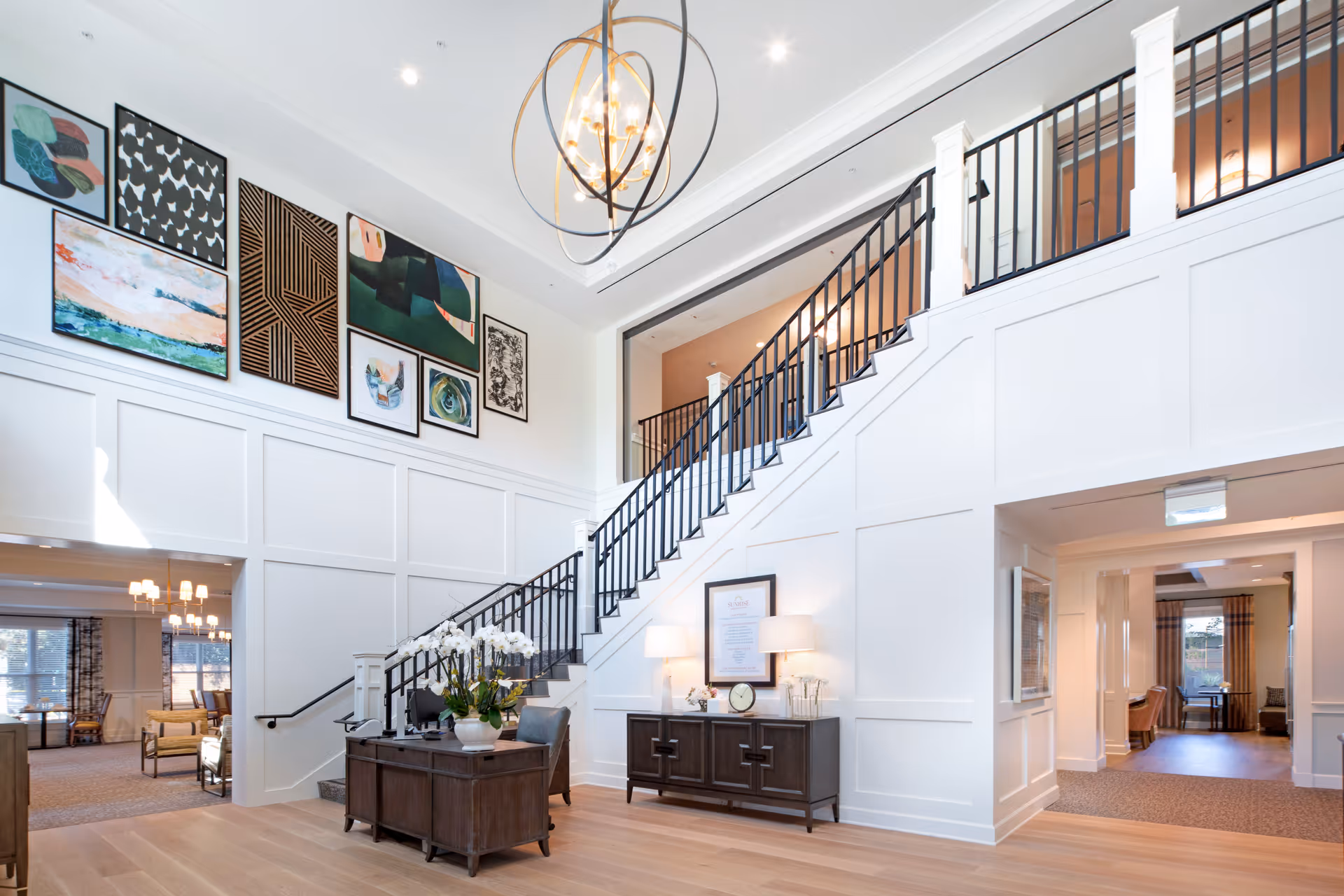 Bright two-story lobby with a staircase, modern chandelier, seating areas, and framed artwork on the walls.