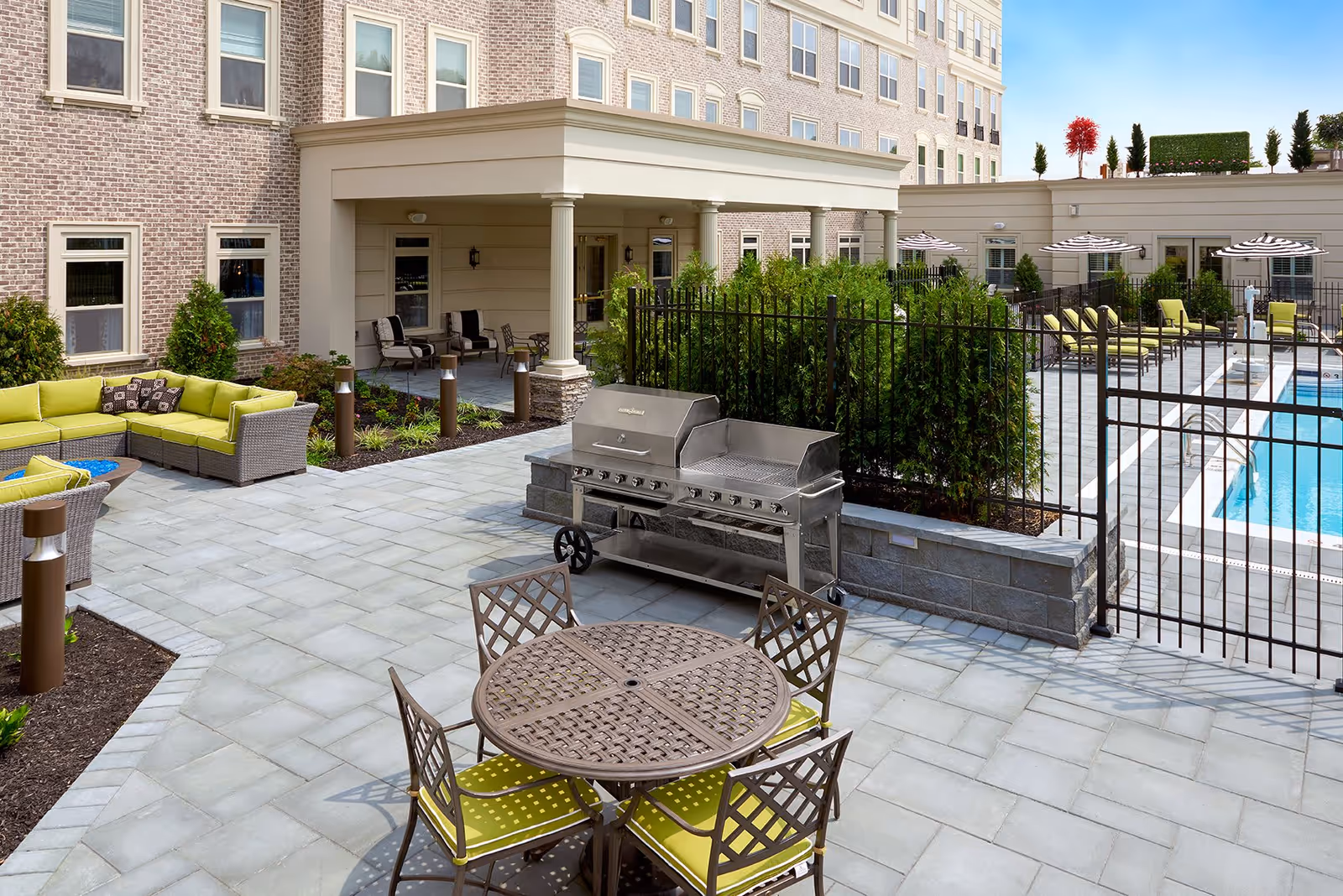 Outdoor patio area at The Bristal at Garden City featuring a round metal table with four chairs with green cushions, a large stainless steel grill, a green cushioned sectional sofa, and a fenced swimming pool with lounge chairs and umbrellas in the background.