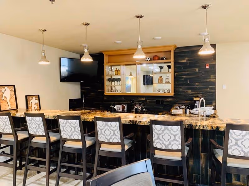 A communal bar area with a long marble countertop, six padded bar stools, pendant lights, a sink and a glass-front cabinet against a dark tiled wall.