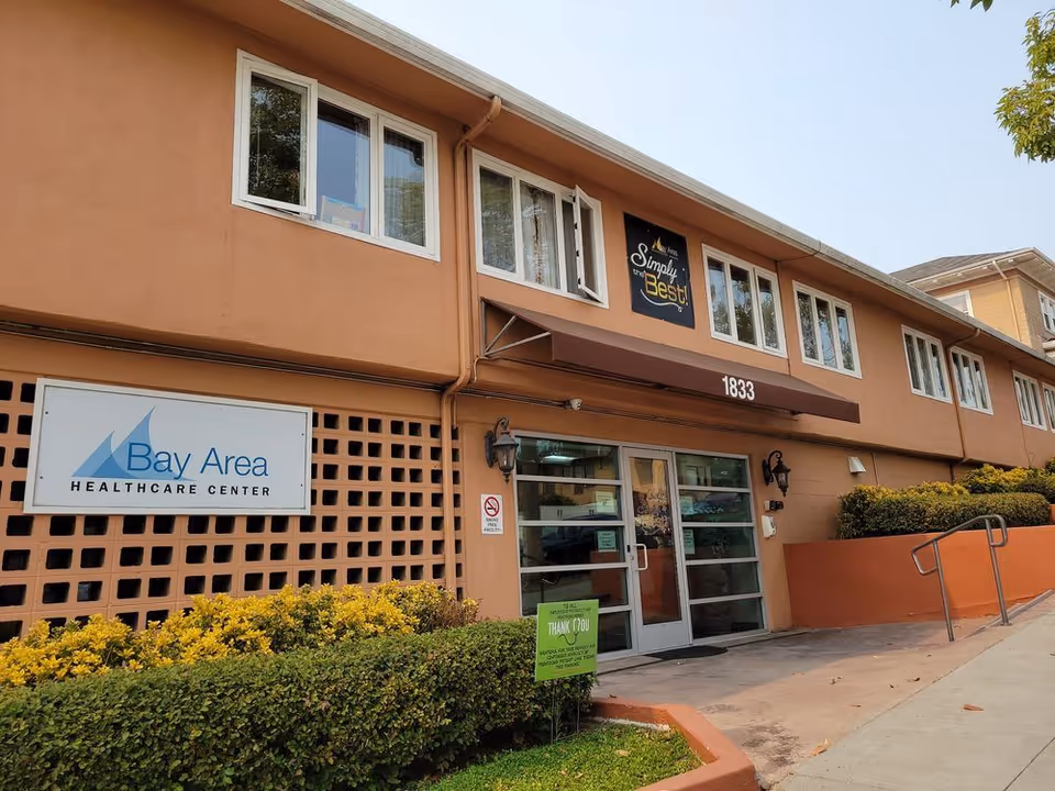 Exterior front entrance of the Bay Area Healthcare Center building with a sign reading 'Bay Area Healthcare Center', address number 1833, and landscaped shrubs.