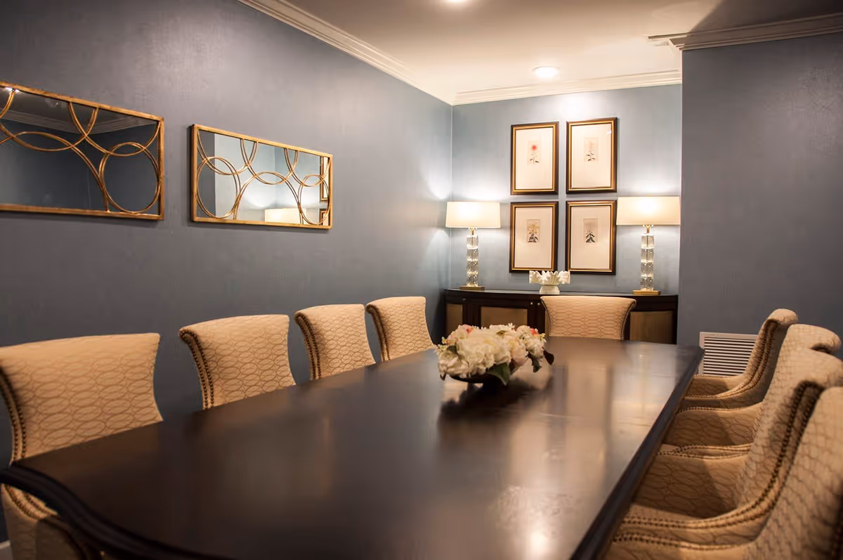 A formal dining room with a long dark wooden table surrounded by beige upholstered chairs. The walls are painted blue and decorated with two rectangular mirrors with gold circular designs on the left and four framed floral artworks on the far wall. Two table lamps with clear glass bases and white shades sit on a dark wooden sideboard beneath the artwork. A floral centerpiece is placed on the dining table.