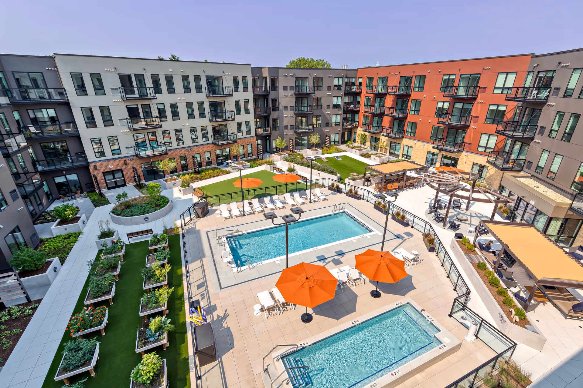 Aerial view of a modern apartment complex courtyard featuring two swimming pools with orange umbrellas and lounge chairs, raised garden beds with plants, green lawn areas, and shaded seating areas with pergolas. The buildings surrounding the courtyard have multiple balconies and large windows.