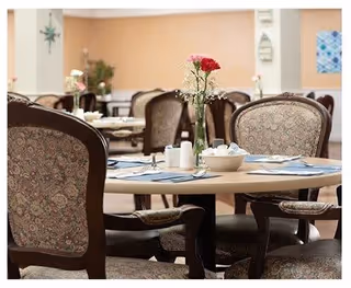 A dining room with round tables set for a meal, featuring floral-patterned upholstered chairs, a vase with red and pink flowers on the table, and soft peach-colored walls with decorative items in the background.