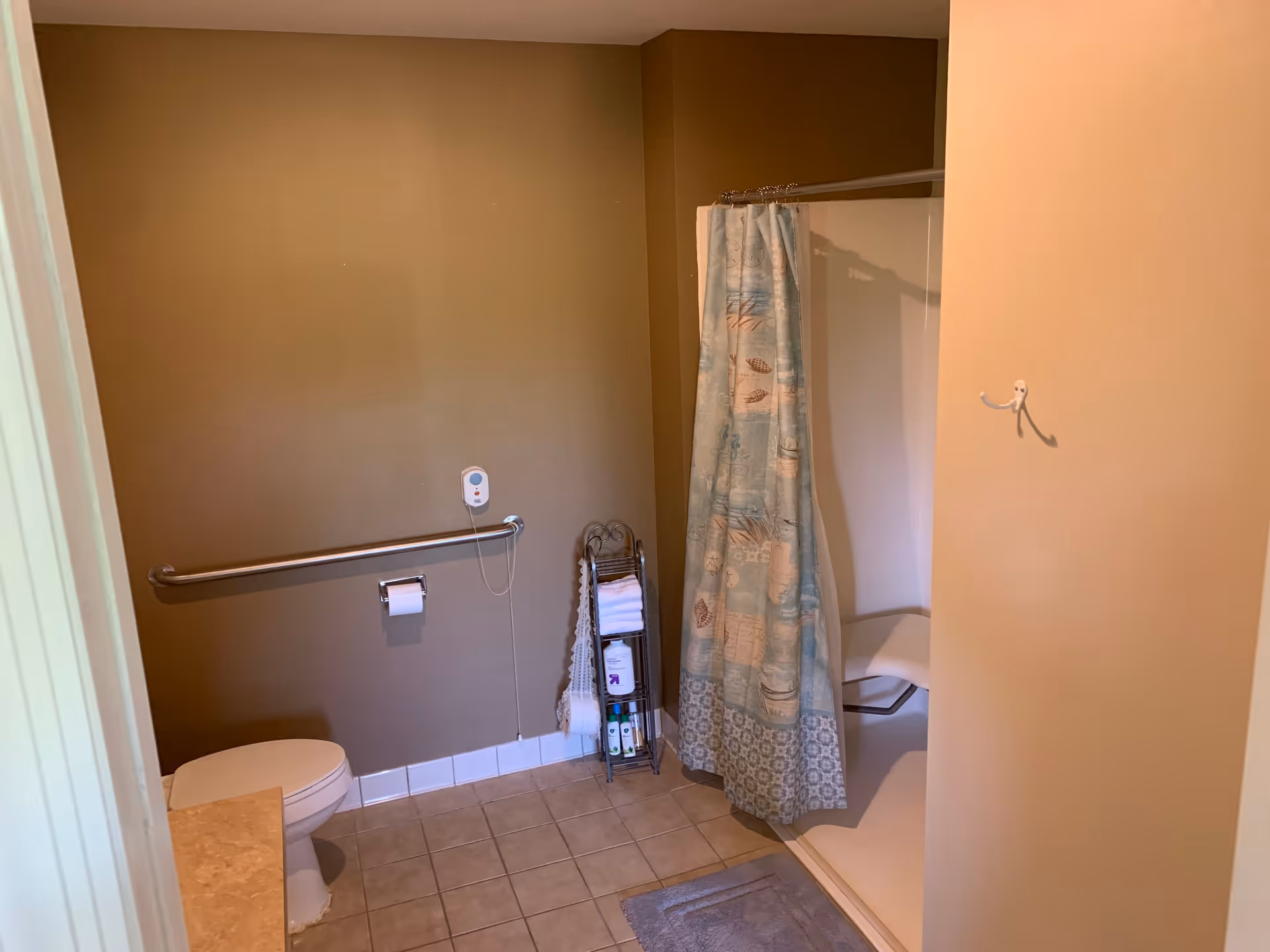 A bathroom with beige walls and tiled floor featuring a toilet with a grab bar and toilet paper holder on the wall. There is a shower area with a patterned shower curtain and a built-in shower seat. A small rack holds towels and toiletries, and a bath mat is placed on the floor in front of the shower.