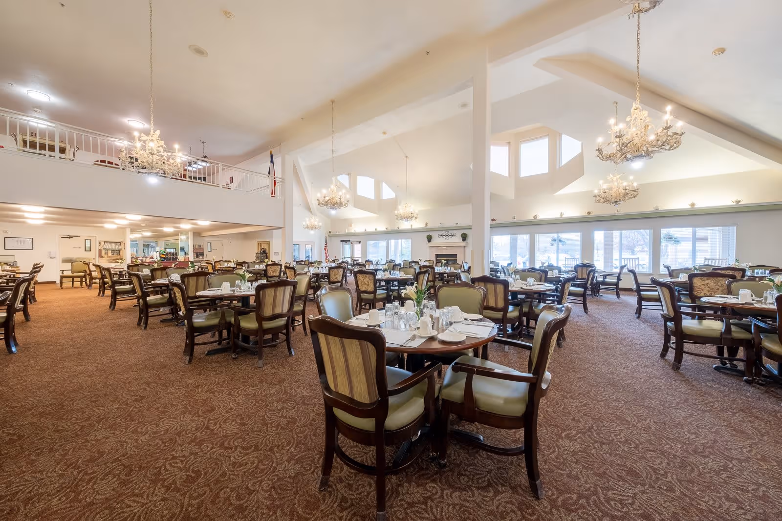Spacious dining room with multiple round tables and cushioned chairs arranged neatly. The room has high ceilings with elegant chandeliers hanging down, large windows letting in natural light, and a carpeted floor with a patterned design. The tables are set with white plates, cups, and silverware, ready for a meal service.