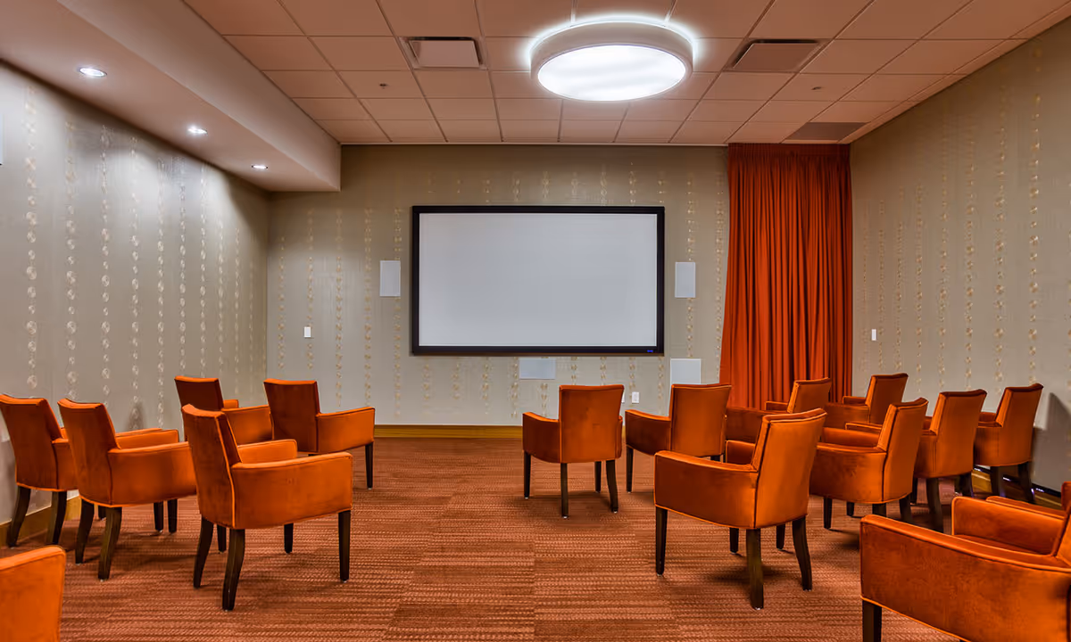 A small theater or presentation room with rows of orange upholstered chairs facing a large blank screen mounted on a beige patterned wall. The room has a carpeted floor matching the chairs and a circular ceiling light above.