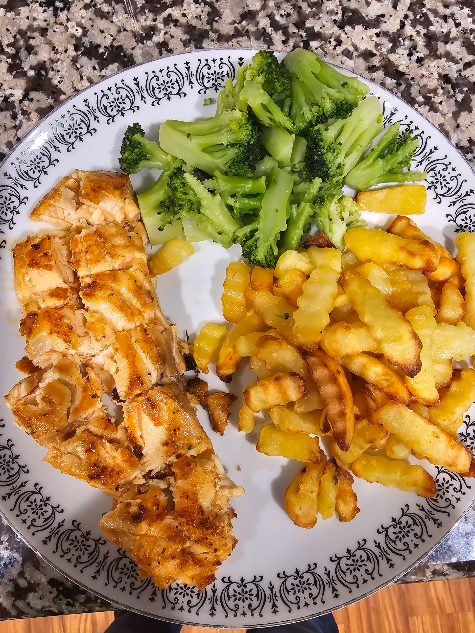 A plate with grilled salmon, steamed broccoli, and crinkle-cut French fries on a granite countertop.