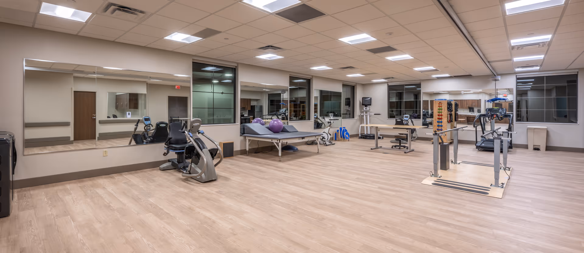 A spacious physical therapy or rehabilitation room with exercise equipment including stationary bikes, parallel bars for walking practice, therapy tables with exercise balls, and various other rehabilitation tools. The room has large mirrors on one wall, light wood flooring, and bright overhead lighting.