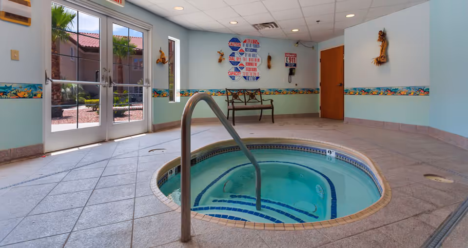 Indoor hot tub with tiled floor and handrail in a room with light blue walls, decorative border, a bench, emergency instructions on the wall, and glass doors leading outside to a garden area with palm trees.