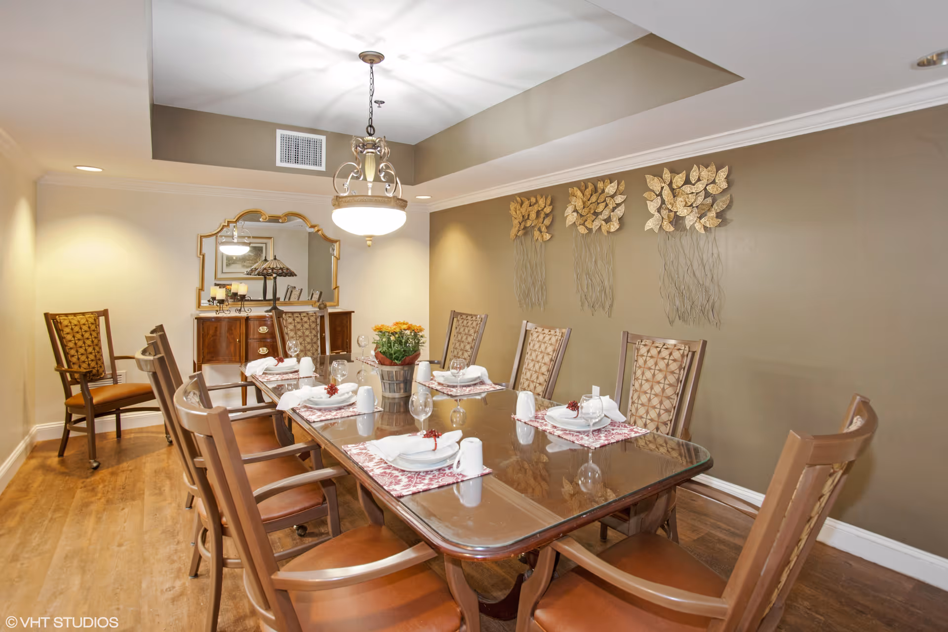 A dining room with a long wooden table set for six people, featuring placemats, plates, napkins, and glasses. The room has wooden chairs with patterned cushions, a decorative wall with three gold leaf wall hangings, a chandelier hanging from the ceiling, and a wooden sideboard with a mirror and decorative items in the background.