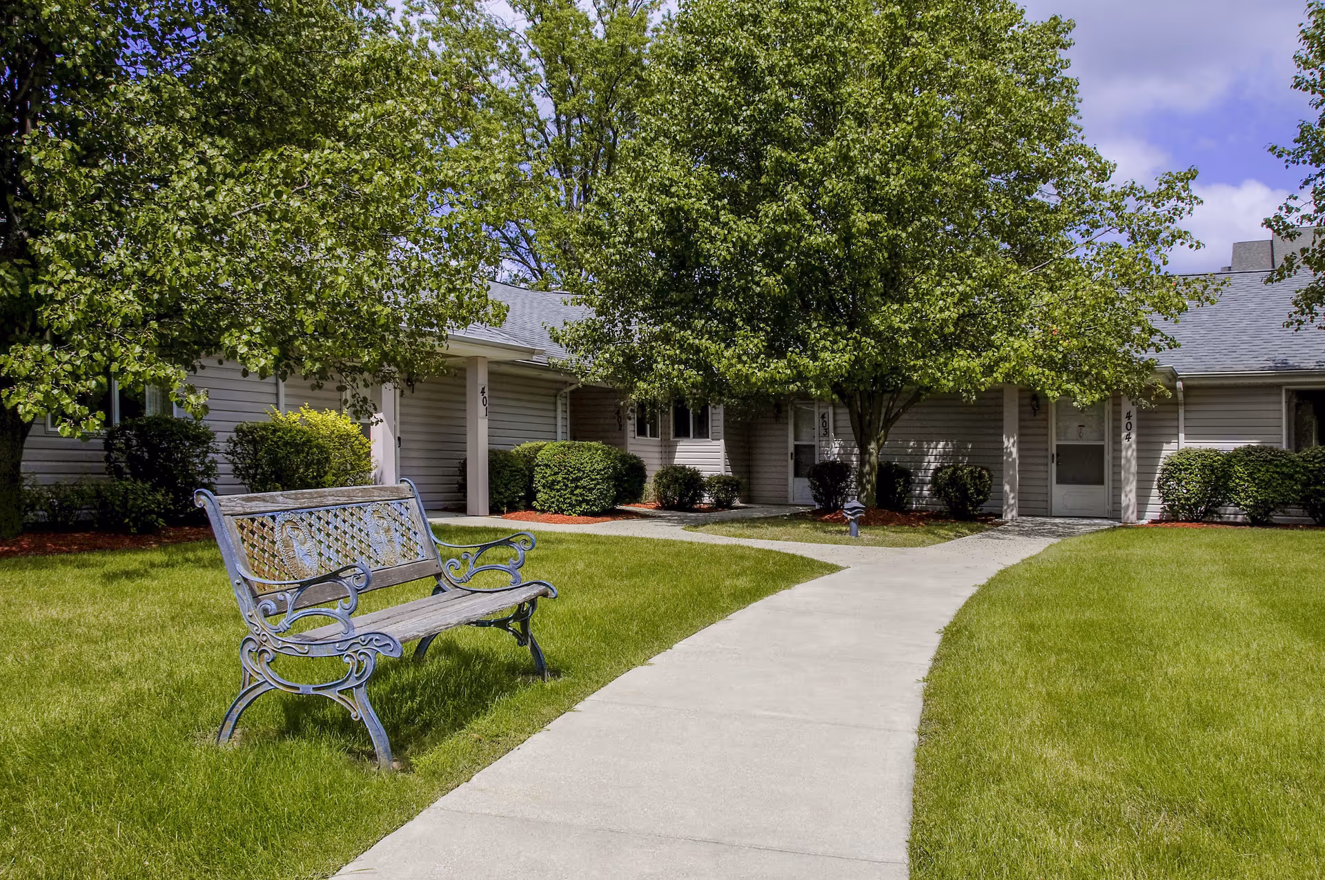 Outdoor view of a senior living facility courtyard with a concrete pathway, green grass, a decorative metal bench, and several leafy trees and bushes in front of single-story building entrances numbered 401, 402, 403, and 404.