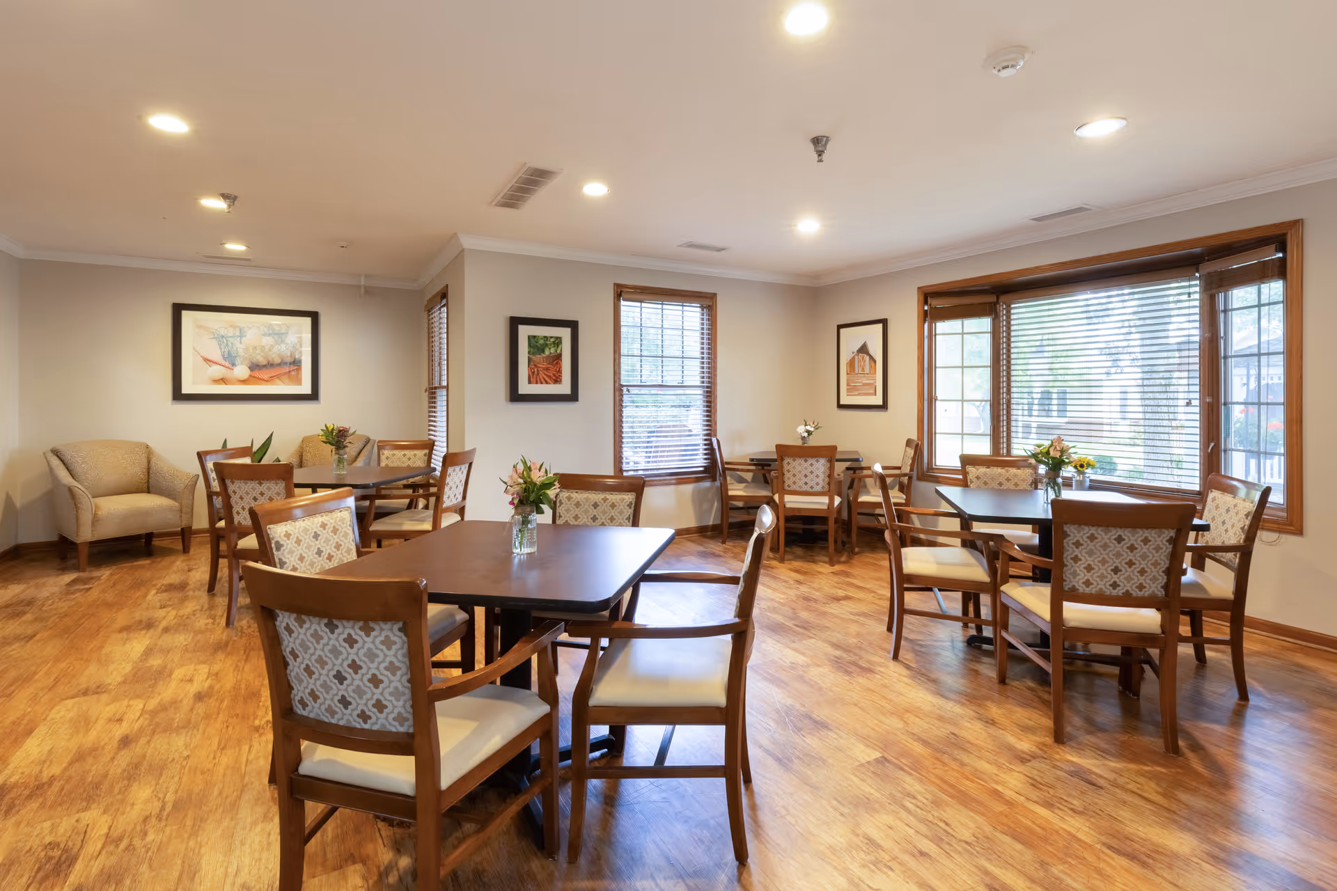 A bright and clean dining room with wooden floors and several square tables surrounded by wooden chairs with patterned cushions. Each table has a small vase with flowers. The room has large windows with wooden blinds, framed artwork on the walls, and recessed ceiling lights.