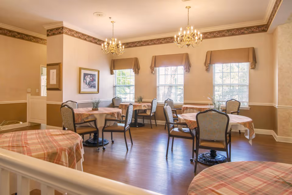 A dining room with several round tables covered with plaid tablecloths and surrounded by chairs. The room has three large windows with valances, two chandeliers hanging from the ceiling, framed artwork on the walls, and wooden flooring.