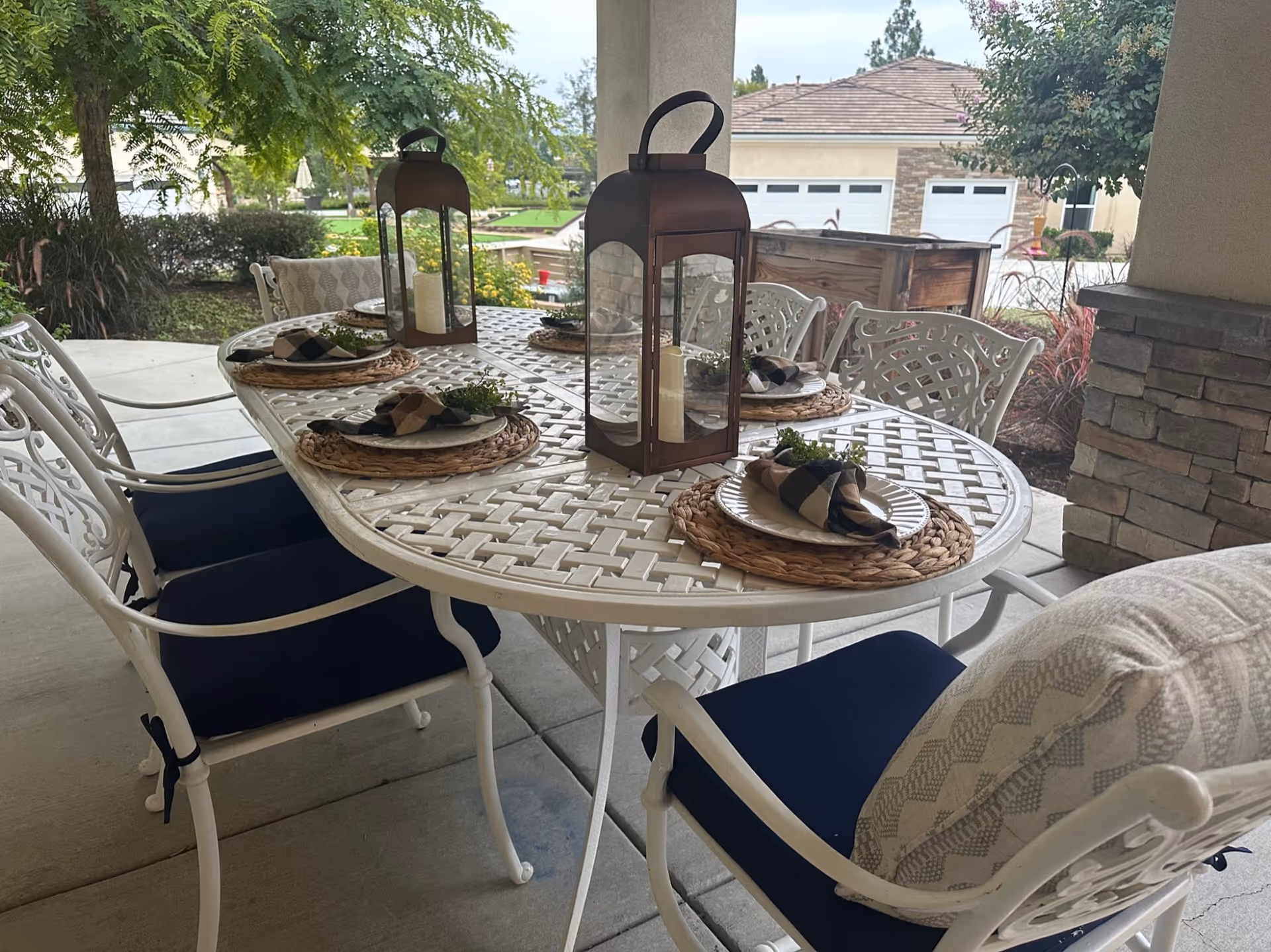 Outdoor patio dining area with a white metal table and six matching chairs with navy blue cushions. The table is set with woven placemats, plates, napkins, and decorative greenery. Two large lanterns with candles are placed on the table. The background shows greenery, trees, and a residential building with garages.