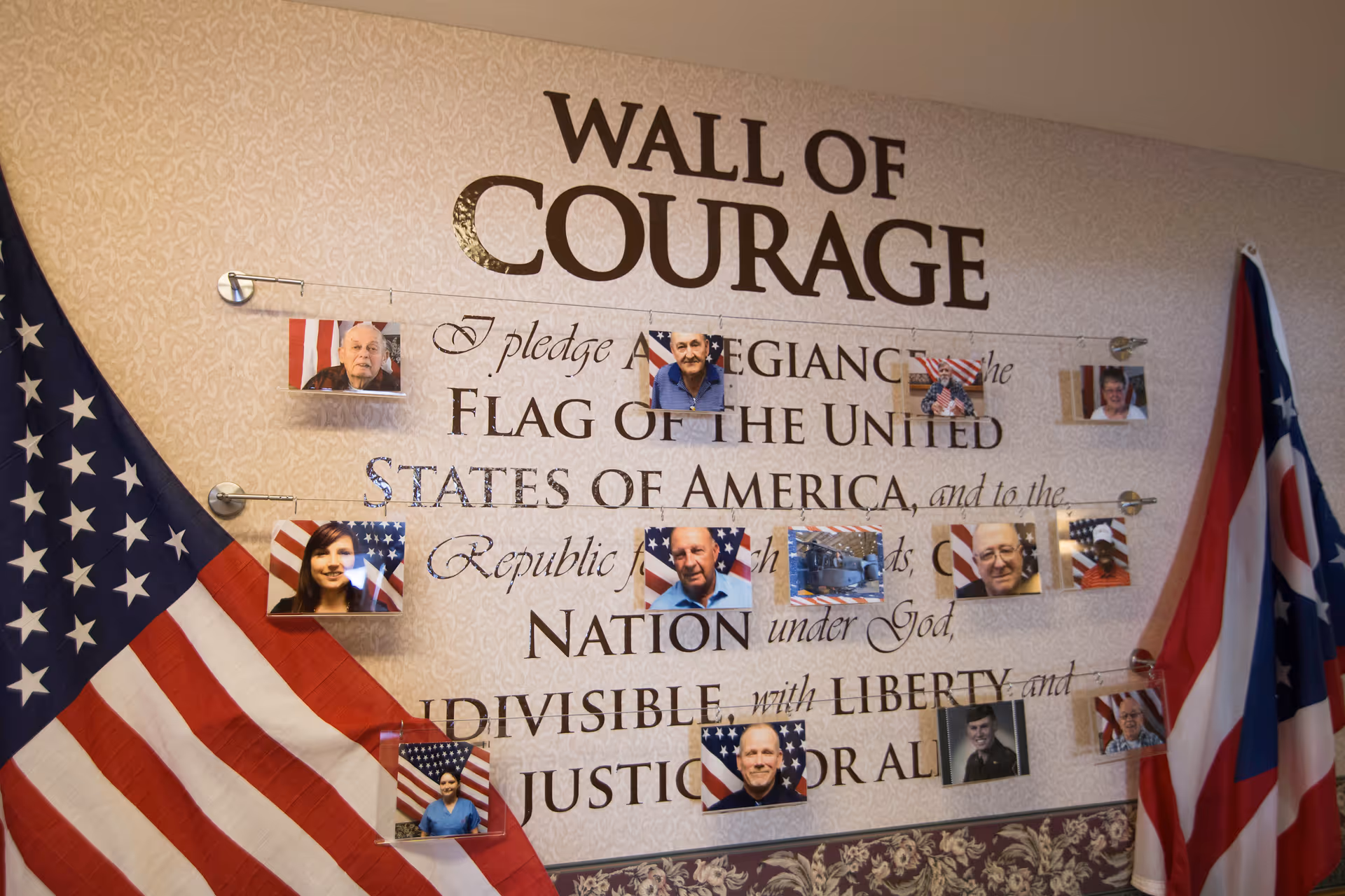 A wall display titled 'Wall of Courage' featuring the Pledge of Allegiance text with photos of various individuals pinned on strings across the wall. Two flags, the United States flag and the Ohio state flag, are draped on either side of the display.