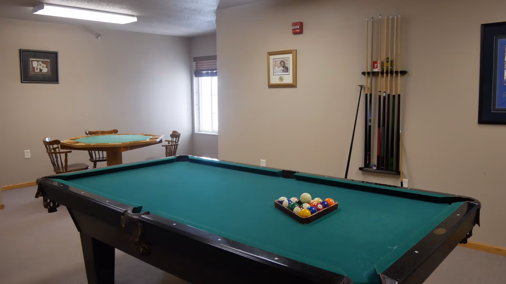 A pool table with racked billiard balls in a game room that also has a card table, chairs, and a cue rack.