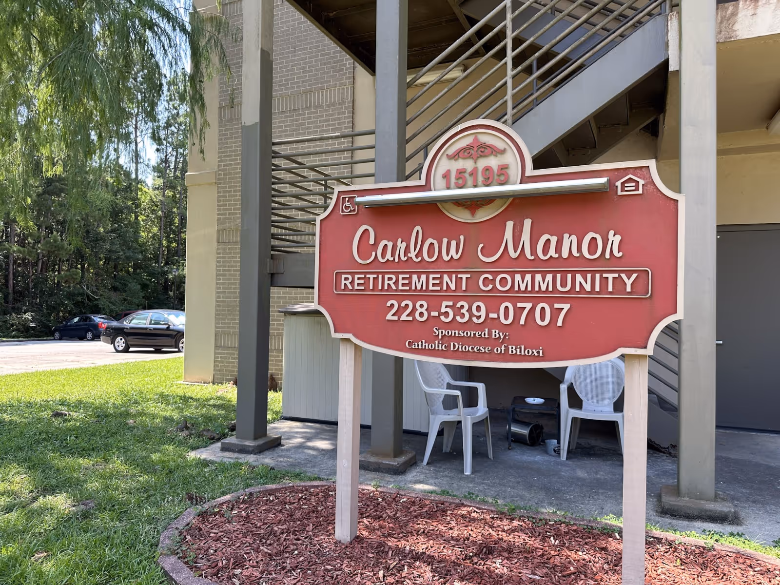 A red Carlow Manor Retirement Community sign stands in front of a building with an exterior staircase and white chairs nearby.