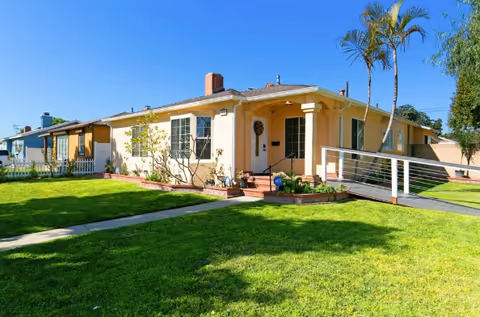 Single-story beige house with a ramp, front yard, and palm trees in a sunny residential setting.