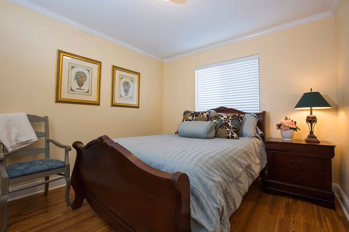 A cozy bedroom with a wooden bed frame and a neatly made bed with multiple decorative pillows. There is a wooden nightstand with a lamp and a small vase of flowers. A wooden chair with a cushion and a folded blanket is placed against the wall. Two framed floral artworks hang on the beige wall, and a window with white blinds is above the bed.