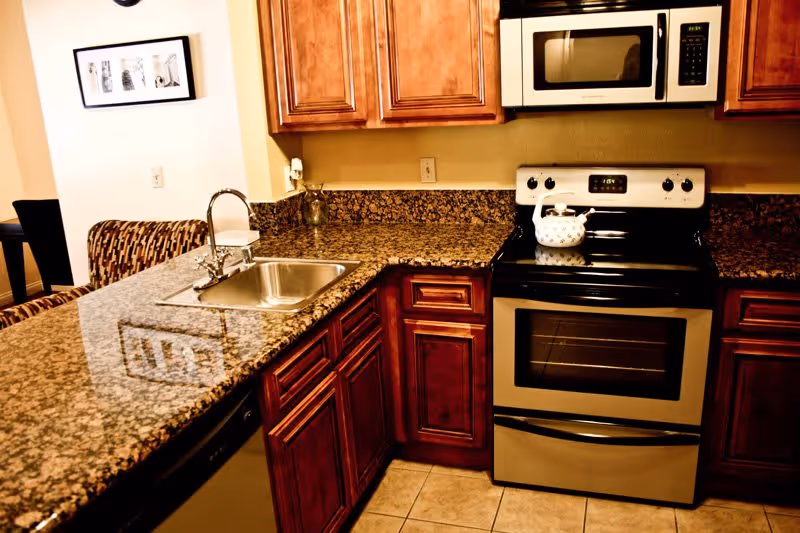 A kitchen with granite countertops, wooden cabinets, a stainless steel sink with a faucet, a stainless steel oven with a white kettle on top, and a microwave above the oven. In the background, part of a dining area with a patterned chair and framed artwork on the wall is visible.