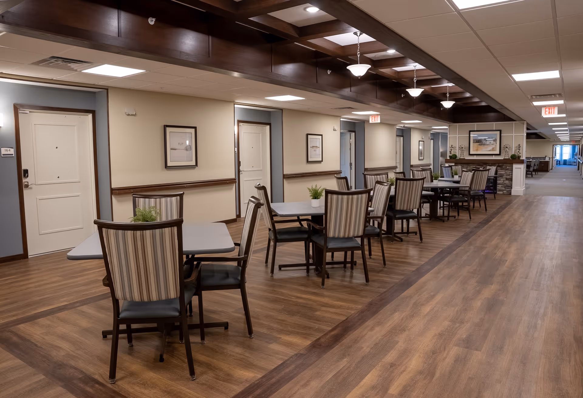 Interior view of a senior living facility hallway with multiple tables and chairs arranged along the wall. The floor is wood-patterned, and the ceiling has recessed lighting and hanging light fixtures. Several white doors with room numbers are visible along the hallway, and framed artwork hangs on the walls. The space is clean and well-lit.