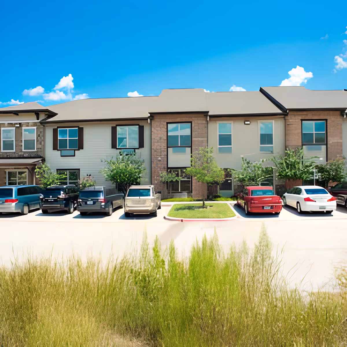Exterior view of a senior living facility building with multiple windows and a parking lot in front. Several cars are parked in the lot, and there are small trees and bushes along the building. The sky is clear and blue with a few clouds.