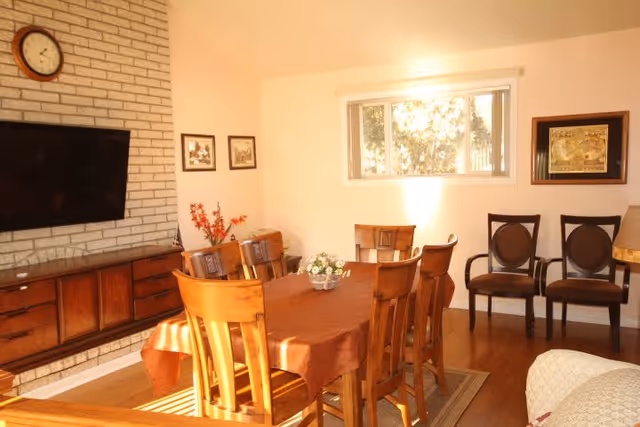 Sunlit dining room with a wooden table and chairs, a wall-mounted TV on a brick accent wall, and additional seating by a window.