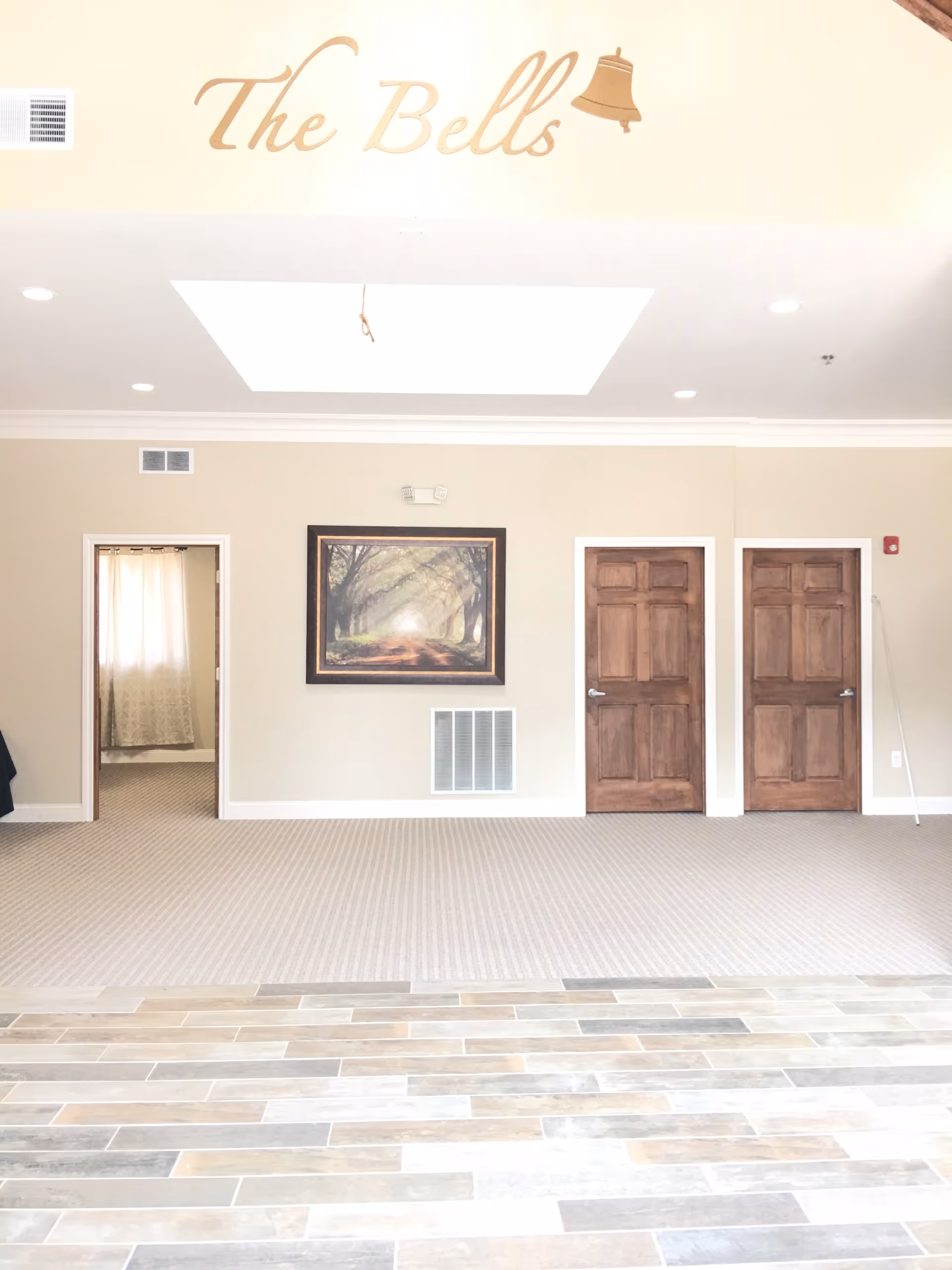 Interior view of a senior living facility named The Bells, showing a beige wall with three wooden doors, a framed picture of a tree-lined path, and a carpeted floor transitioning to a tiled floor in the foreground. The facility name 'The Bells' is displayed in gold lettering with a bell icon above the doors.