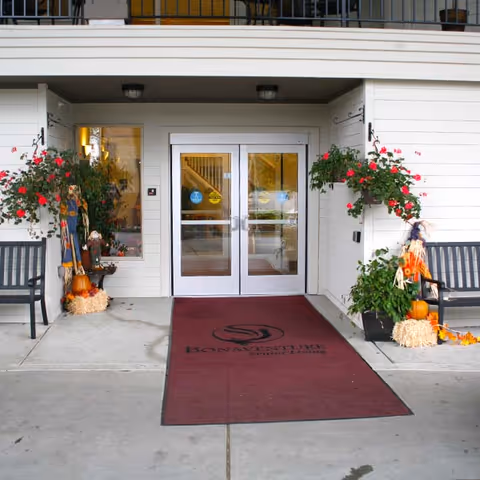 Entrance to a building with double glass doors, flanked by windows and decorated with hanging flower baskets and autumn-themed decorations including pumpkins and hay bales. A maroon mat with the Bonaventure logo and text is placed in front of the doors, with benches on either side.