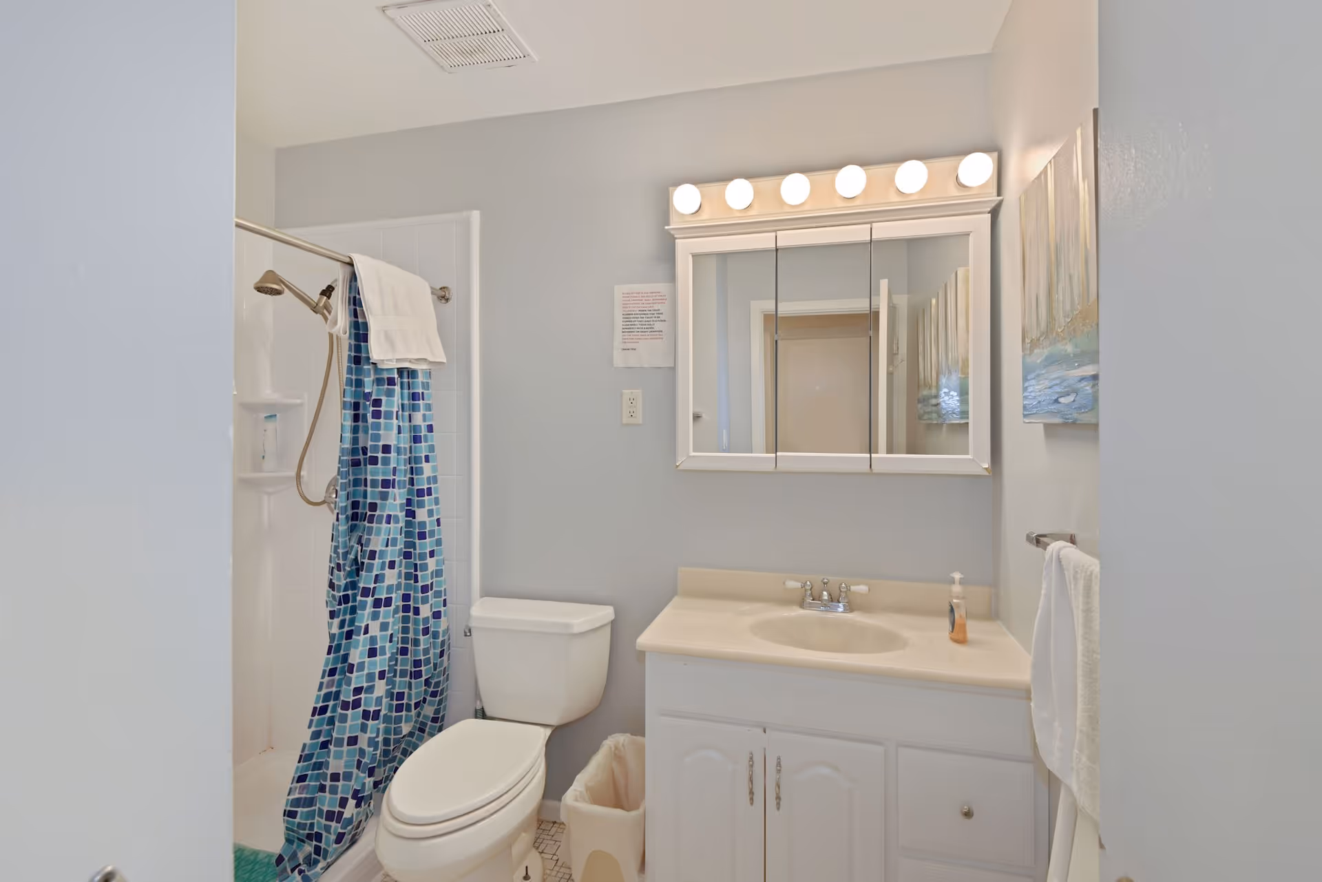 A clean bathroom with a white toilet, a vanity with a sink and a mirror cabinet above it with lights. There is a shower with a blue and white patterned curtain and a towel hanging on the shower rod. A white towel hangs on a towel rack on the right wall, and a small trash bin is placed next to the toilet. The walls are painted light gray and there is a piece of artwork on the right wall.