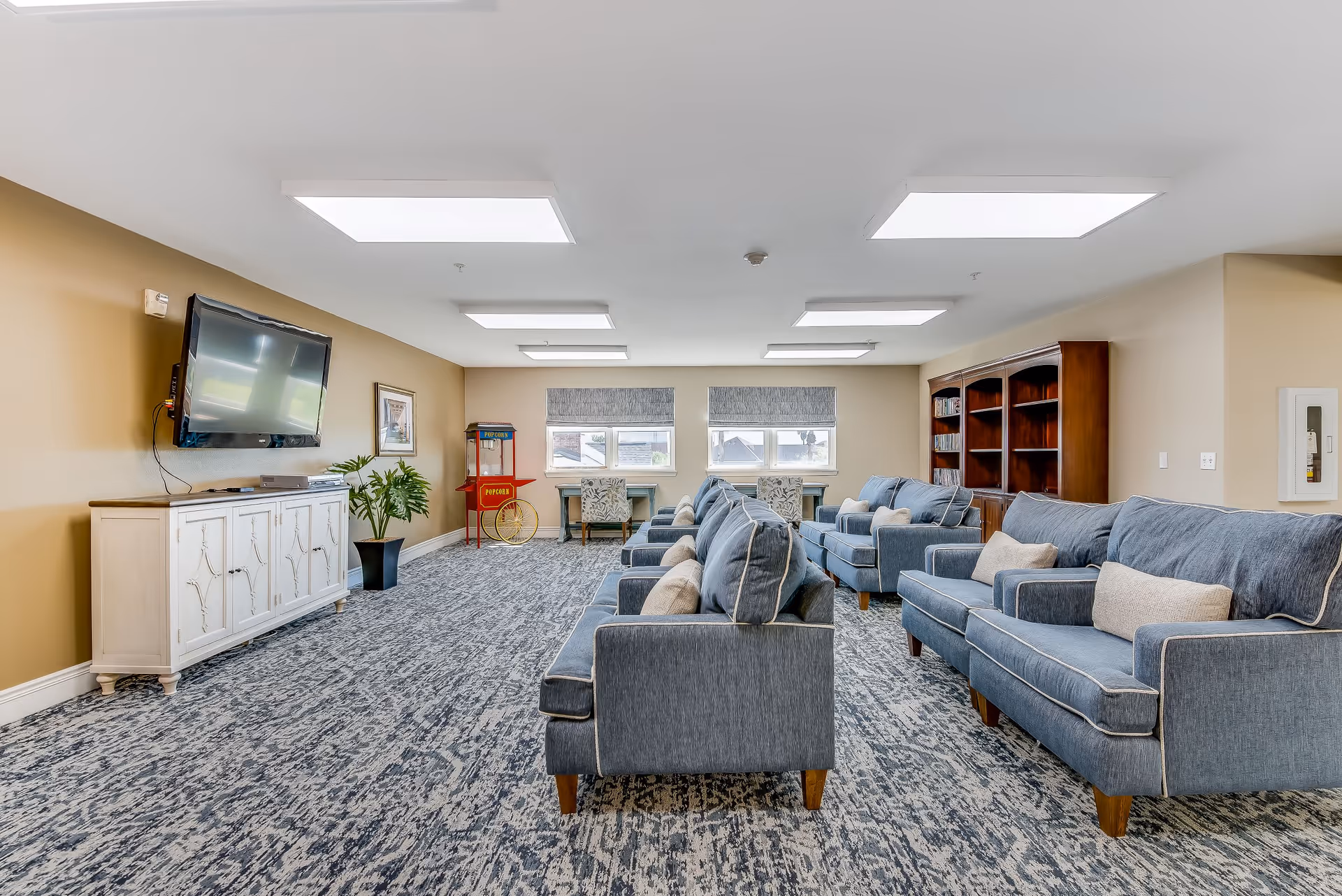 A spacious senior living common area with multiple blue upholstered sofas arranged in two rows facing a wall-mounted flat-screen TV. The room has a patterned carpet, beige walls, and ceiling lights. There is a white cabinet under the TV, a potted plant, a popcorn machine in the corner, two windows with Roman shades, two desks with chairs, and a large wooden bookshelf on the right side.