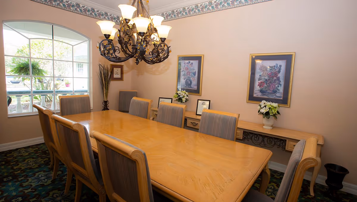 A dining room with a large wooden table surrounded by eight cushioned chairs. A decorative chandelier with multiple lights hangs above the table. The room has a large window with a view of greenery outside, floral paintings on the wall, and a console table with flower arrangements and framed certificates.