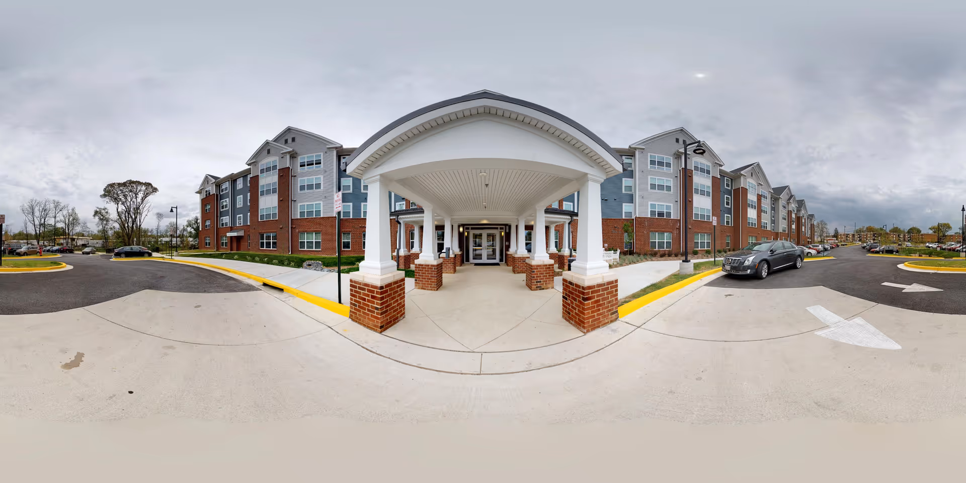 Front exterior view of The Belnor Senior Residences building with a covered entrance supported by white columns and brick bases. The building is multi-story with a combination of brick and siding, surrounded by a parking lot with cars and street lamps under a cloudy sky.