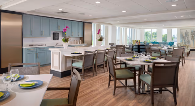 Bright communal dining room with multiple set tables, chairs, a central island and blue kitchenette cabinets.