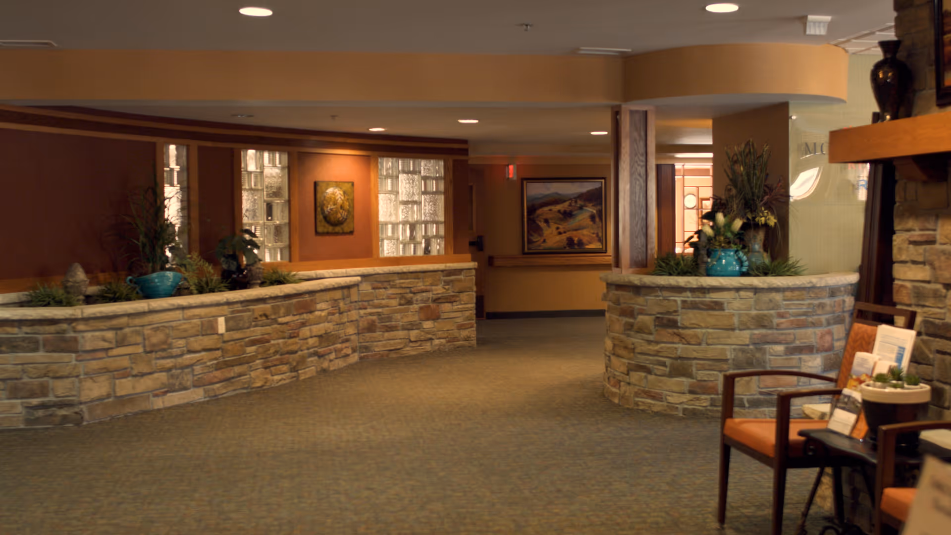 A warm interior lobby with curved stone planter walls, potted plants, artwork, and seating.