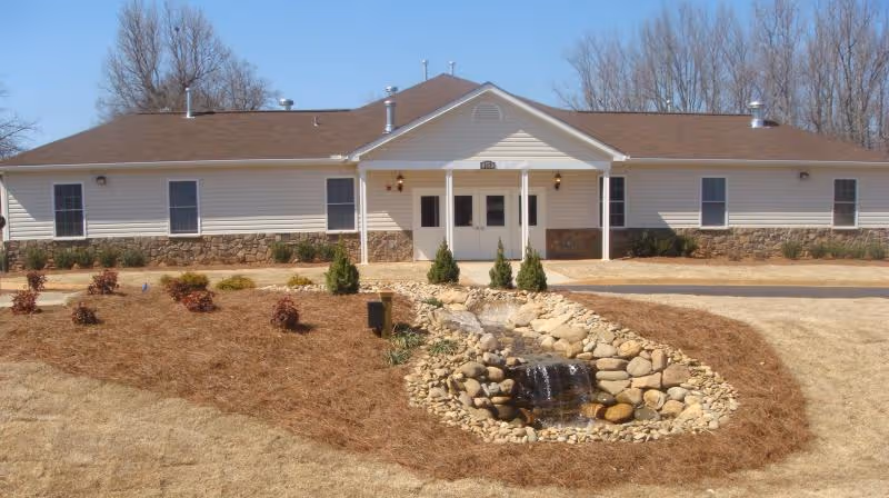 Single-story light-colored building front with a central covered entrance, landscaped beds, and a small rock waterfall in the lawn.
