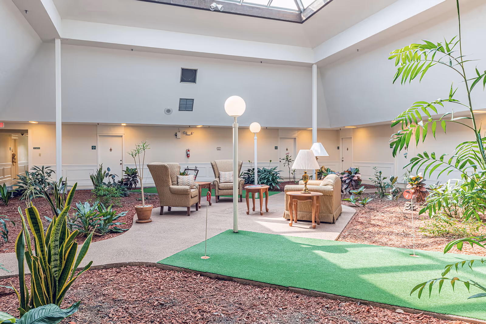 Atrium-style indoor lounge with armchairs, side tables, lamps, plants and a small putting green under a skylight.
