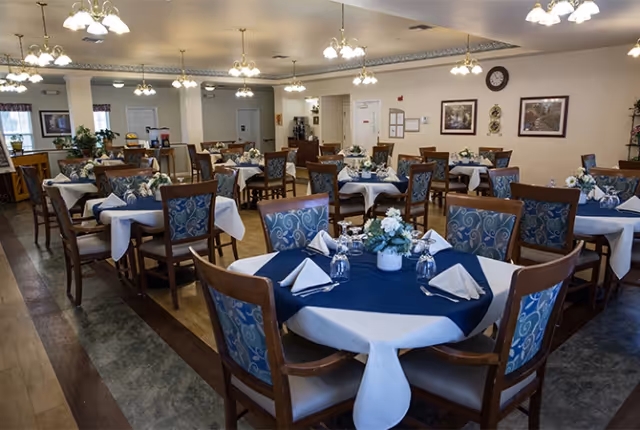 A dining room with multiple round tables covered with white and blue tablecloths, each set with folded napkins, glasses, and floral centerpieces. The room has wooden chairs with blue patterned cushions, hardwood flooring, and several ceiling light fixtures. The walls are decorated with framed pictures and a clock.