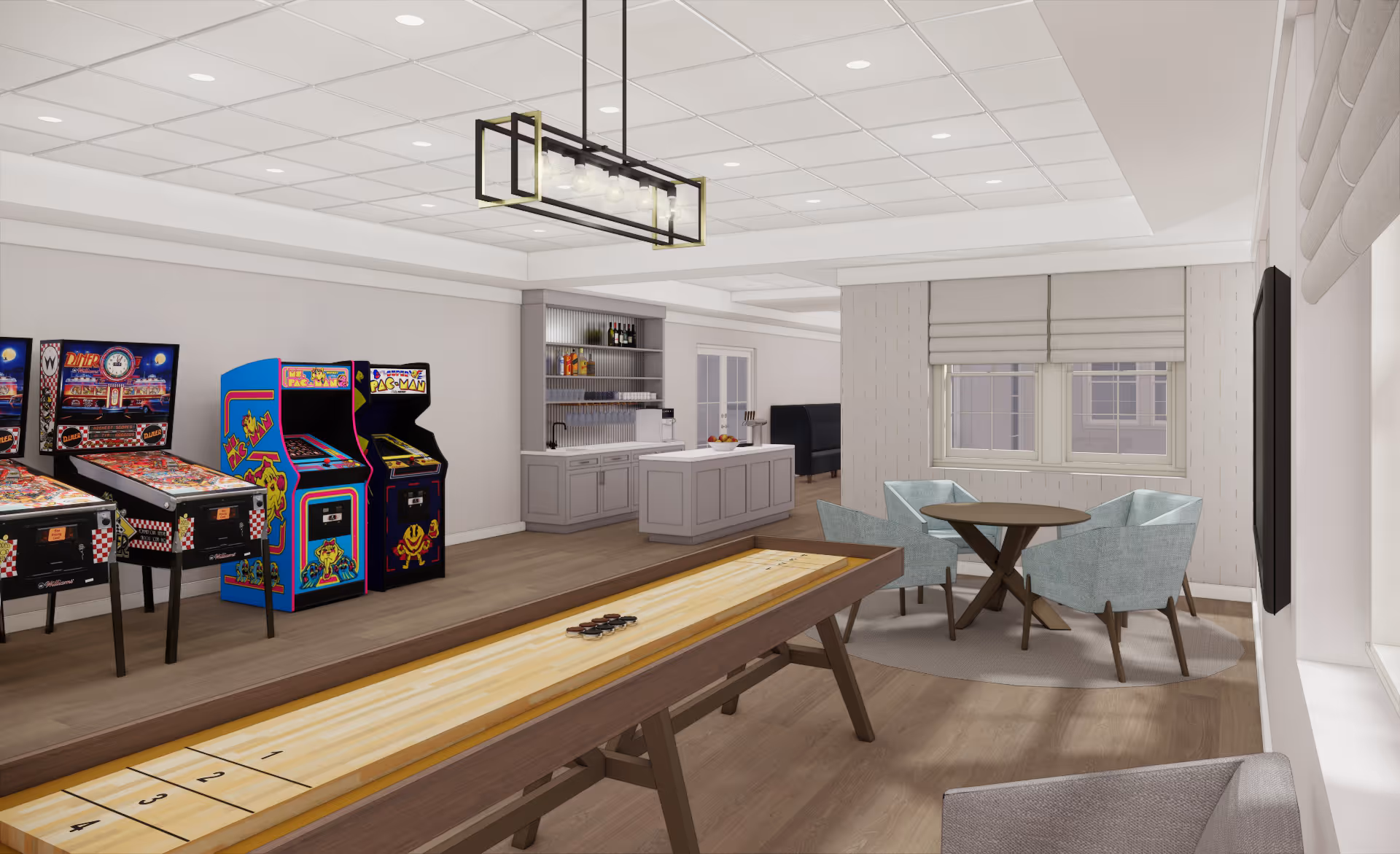 A recreational room featuring two pinball machines and two classic arcade game machines against the wall, a shuffleboard table in the center, a small round table with four light blue chairs on a circular rug near two windows with blinds, and a kitchenette area with cabinets, shelves, and a countertop in the background.