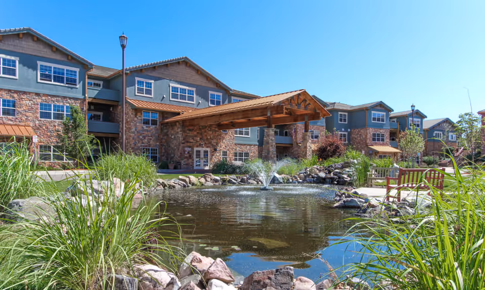 Exterior view of Keystone Place At Legacy Ridge senior living facility featuring a stone and wood building with multiple windows, a covered entrance, a pond with a water fountain, surrounding rocks, green plants, benches, and a clear blue sky.