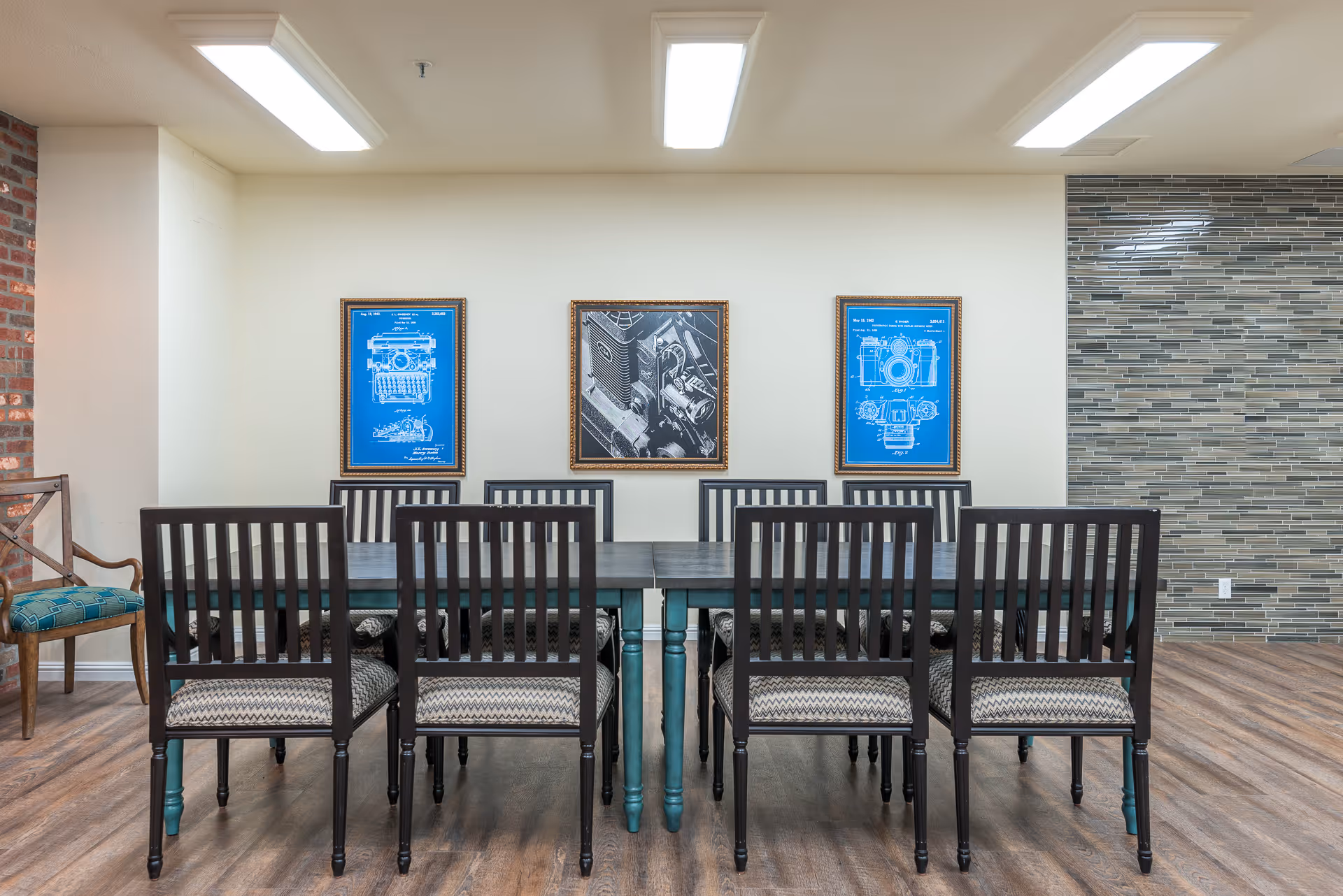 Long rectangular dining table with eight wooden chairs in a bright room with three framed blueprint-style prints on the wall.