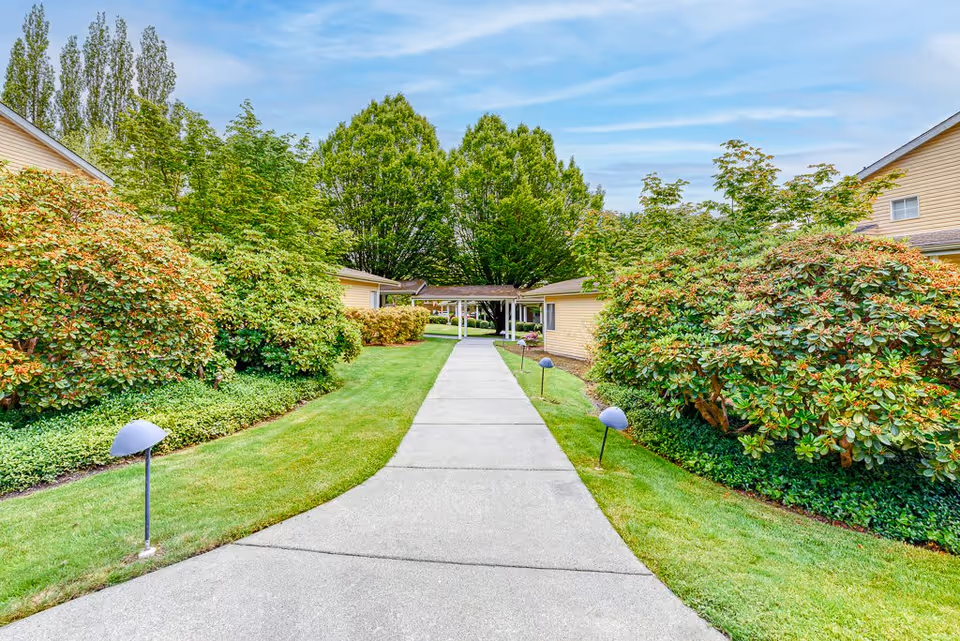 A paved walkway flanked by green grass and well-maintained bushes leads to a covered entrance between two single-story buildings with beige siding. Tall trees are visible in the background under a partly cloudy sky.
