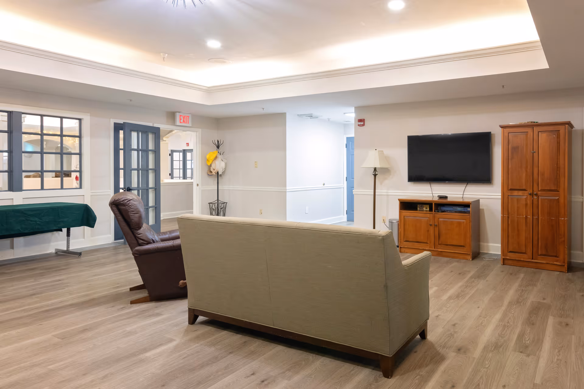 Spacious common living room with a sofa and recliner facing a wall-mounted TV above a wooden media cabinet, with an armoire and table in the background.